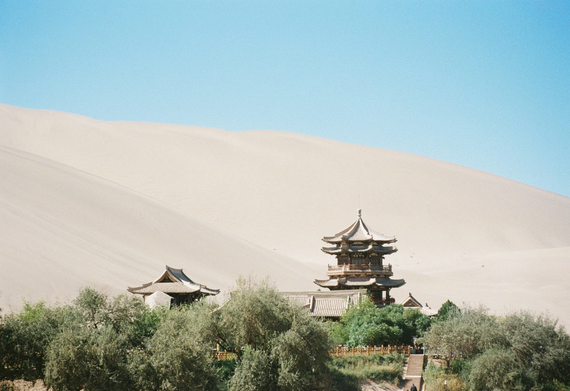 Traditional pavilion and green oasis trees below the bare dunes of Mingsha Mountain in Dunhuang