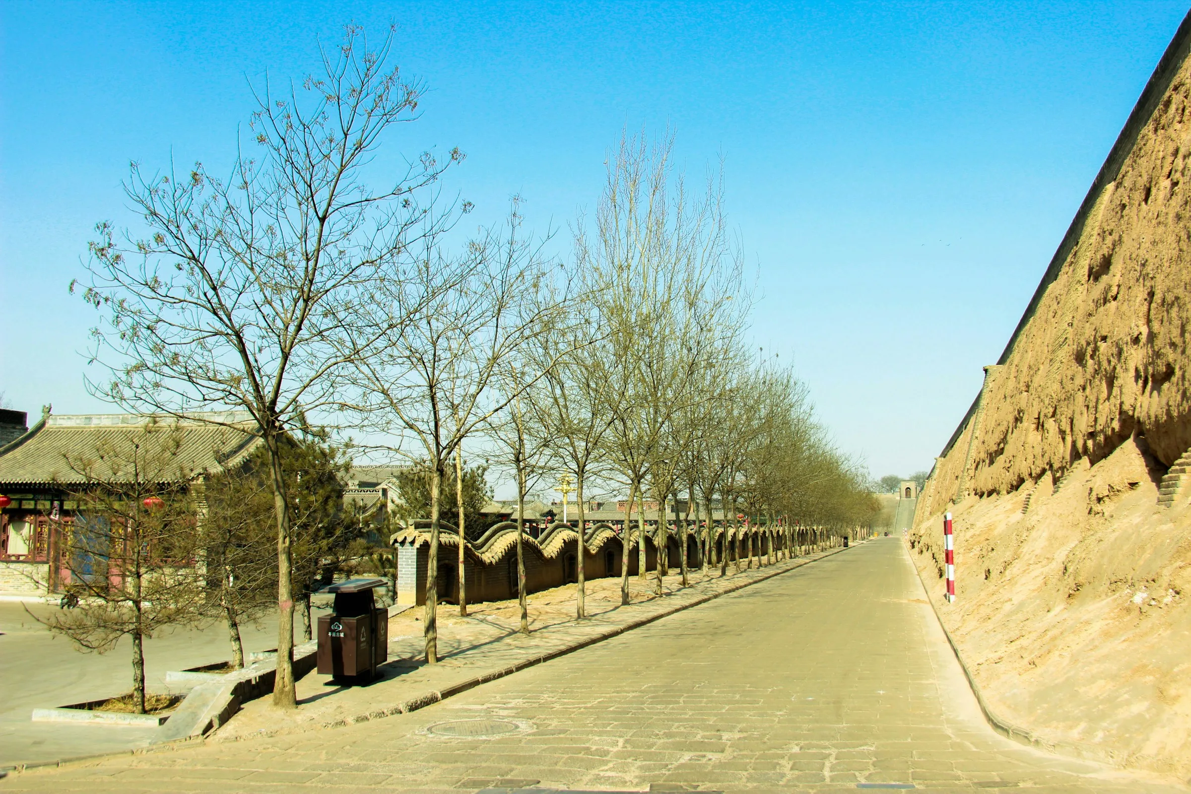 Tree-lined path running beside the tall textured outer face of Pingyao's ancient city wall